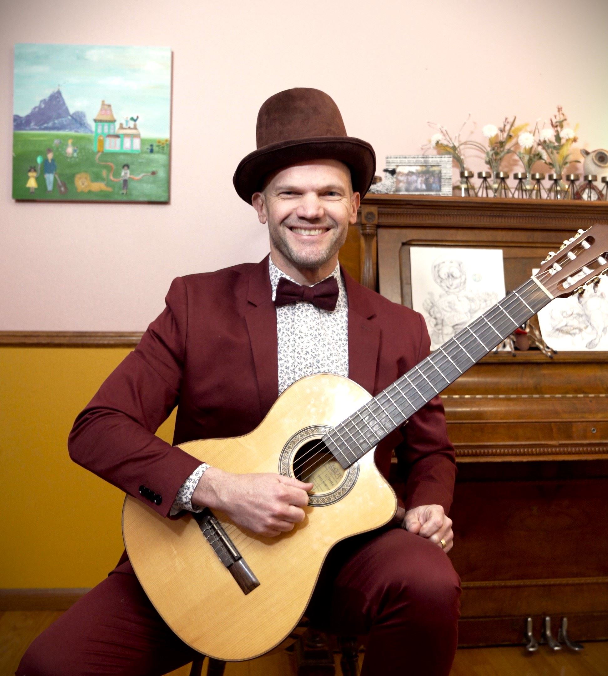 Smiling performer Duke Otherwise in burgundy top hat and suit, sitting near piano with guitar. 